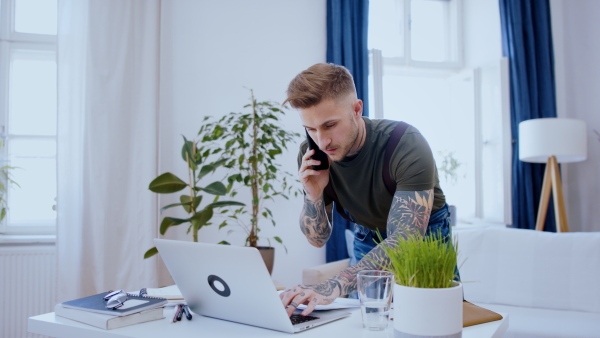 A man with smartphone and laptop indoors at desk in home office, working.