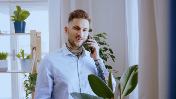 Young man with smartphone standing indoors in home office, making phone call.