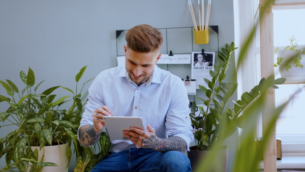 Young man with tablet sitting indoors at desk in home office, working.