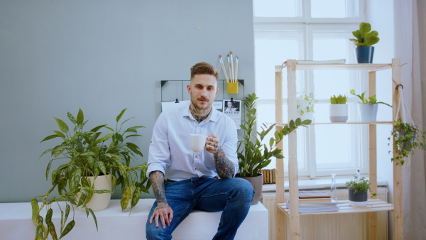 Portrait of man with coffee sitting indoors at desk in home office, looking at camera.