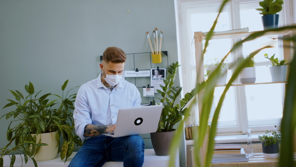 Young man with face mask and laptop sitting indoors in home office, working.