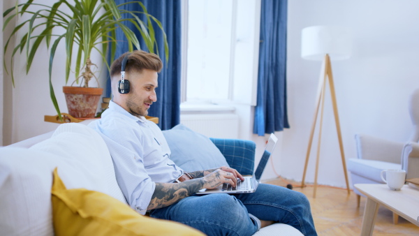 Side view of man with laptop working indoors on sofa in home office, video call concept.