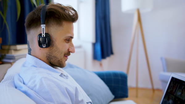 Side view of man with laptop working indoors on sofa in home office, video call concept.