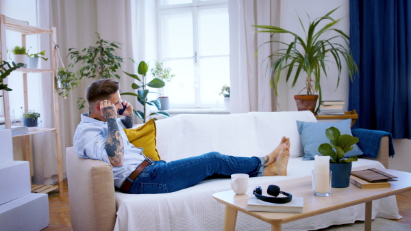 Young man with smartphone sitting indoors on sofa in home office, making phone call.