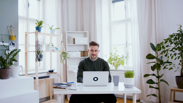 Young man with laptop sitting indoors at desk in home office, working.