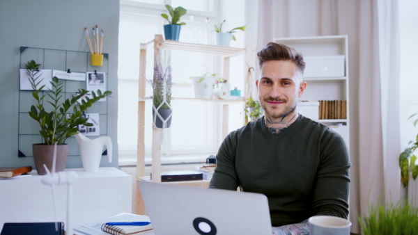 Portrait of man with laptop working indoors at desk in home office, looking at camera.