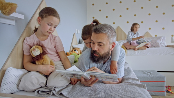 A father with three daughters indoors at home, reading a book.