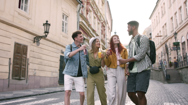 A front view of group of happy young people outdoors on street on town trip, walking with drinks.