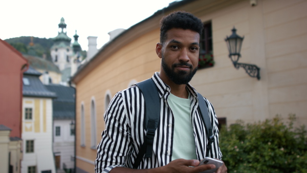 A young man outdoors on trip in town, using online map on smartphone and looking at camera.