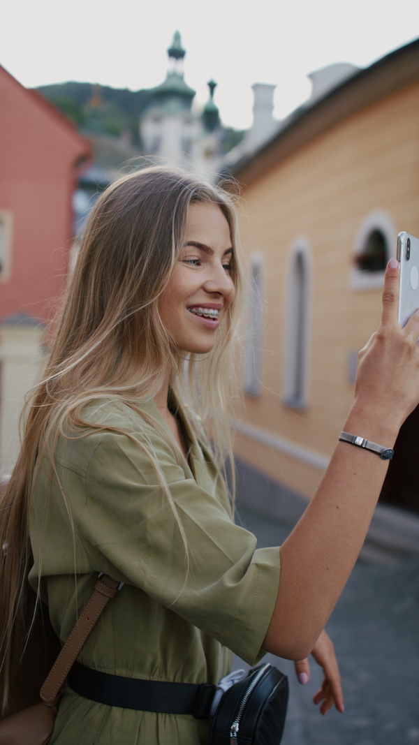 A vertical footage of young woman tourist outdoors on trip in town, taking selfie with smartphone.