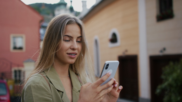 Happy young woman tourist outdoors on trip in town, using online map on smartphone and looking at camera.