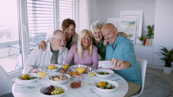 A group of senior friends having party indoors, taking selfie when eating at the table.