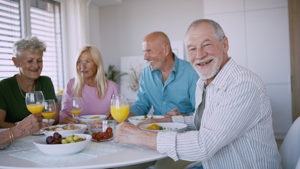 A senior man with friends having party indoors, eating at the table and looking at camera.