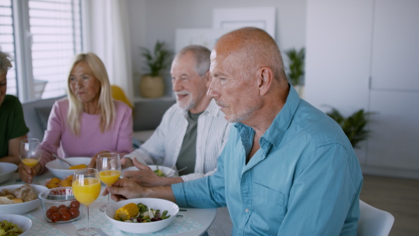 A senior man with friends having party indoors, eating at the table and looking at camera.