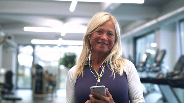 A front view of senior woman with earphones and smartphone in gym resting after doing exercise.