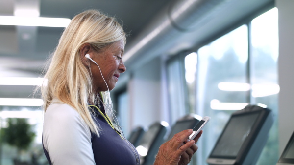 A side view of senior woman with earphones and smartphone in gym resting after doing exercise.