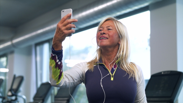 A senior woman with earphones and smartphone in gym resting after doing exercise, taking selfie.