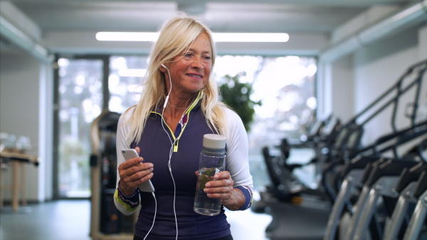 A front view of senior woman with earphones and smartphone in gym resting after doing exercise.