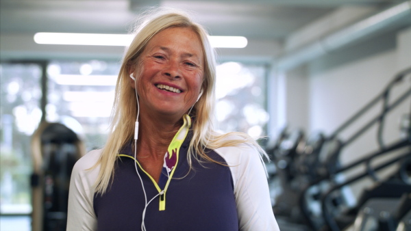 A front view of senior woman with earphones in gym resting after doing exercise.