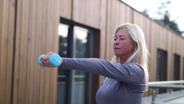 A senior woman with dumbbells outdoors doing exercise on terrace.