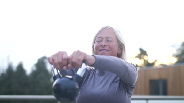 A senior woman with earphones and kettlebell outdoors doing exercise on terrace.