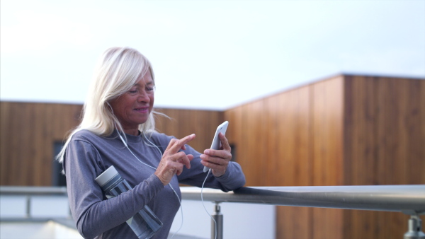 A senior woman with earphones and smartphone outdoors resting after exercise.