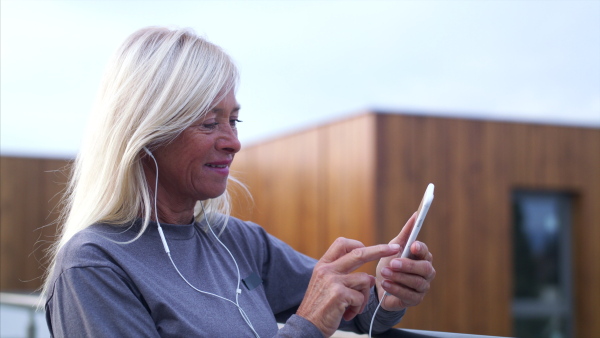 A senior woman with earphones and smartphone outdoors resting after exercise.