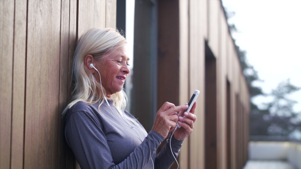 A senior woman with earphones and smartphone outdoors resting after exercise.