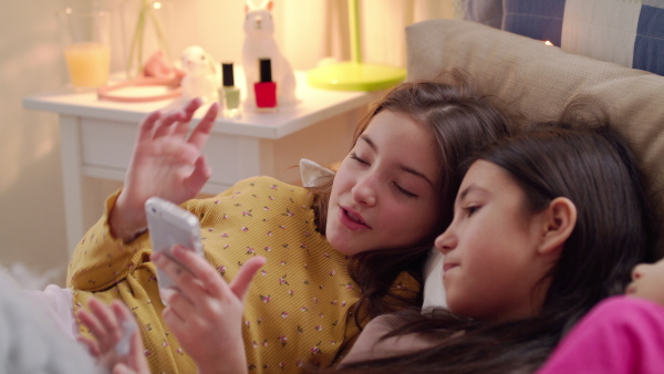 Group of small girls friends lying on bed on slumber party, using smartphone.