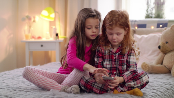 Front view of small girls friends lying on bed on slumber party, using smartphone.