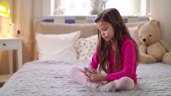 Portrait of small girl with smartphone sitting on bed, danger of addiction concept. Slow motion.