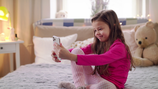 Portrait of happy small girl with smartphone sitting on bed, taking selfie.