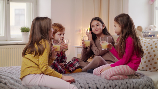 Group of small girls friends in pajamas drinking juice on bed on slumber party.