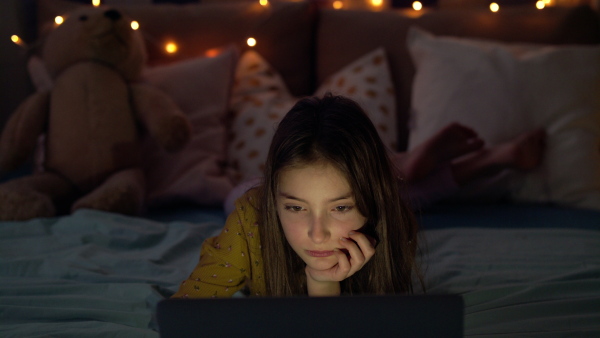 Front view of small girl with laptop computer on bed watching film at night.