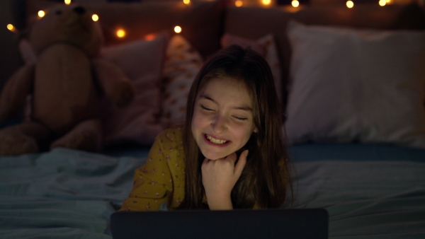 Front view of small girl with laptop computer on bed watching film at night, laughing.