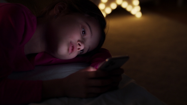 Close-up portrait of small girl lying on bed at night, using smartphone.