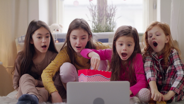 Front view of group of surprised small girls friends watching film on bed on slumber party, eating popcorn.