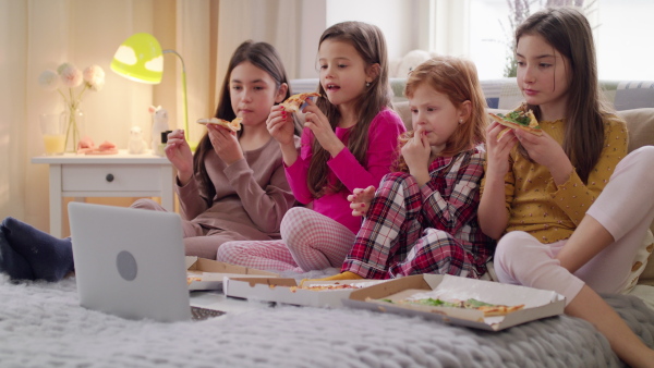 Front view of group of small girls friends watching film on bed on slumber party, eating pizza.