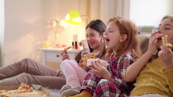 Group of small laughing girls friends watching film on bed on slumber party, eating pizza.