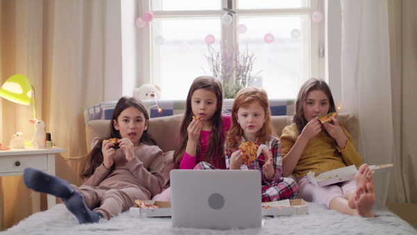 Front view of group of small girls friends watching film on bed on slumber party, eating pizza.