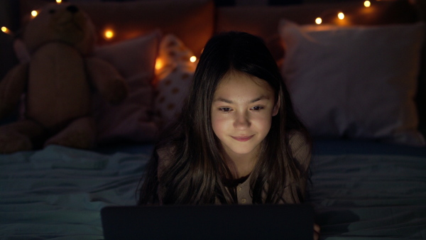 Front view of small girl with laptop computer on bed watching film at night.