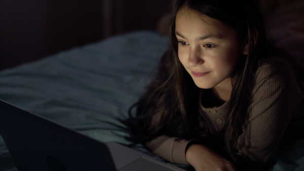 Small girl with laptop computer on bed watching film at night.