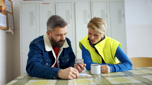 Man and woman worker sitting indoors by lockers, using smartphone during break.