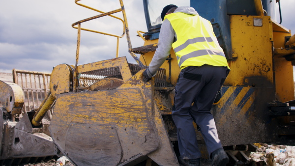 Worker getting in excavator on landfill, waste management and environmental concept.