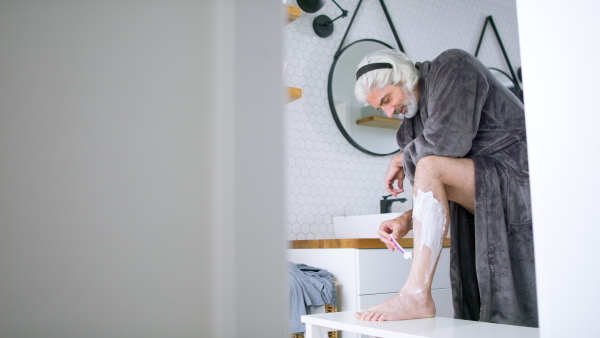 A mature man with grey hair shaving his legs in the bathroom, getting ready for day.