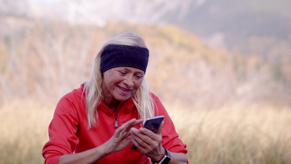 An active senior woman hiker standing outdoors in nature, using smartphone.