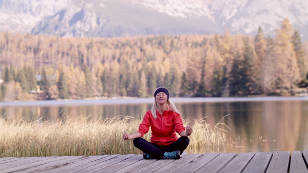 Front view of senior woman hiker sitting outdoors by lake in nature, doing yoga exercise.