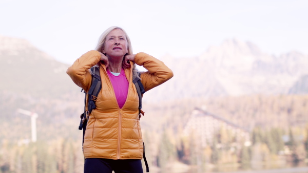 Front view of active senior woman hiker standing outdoors in nature, zipping up a jacket.