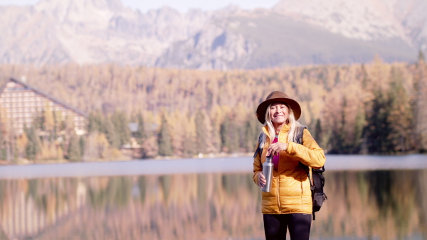 Active senior woman hiker standing by lake outdoors in nature, drinking. Slow motion.