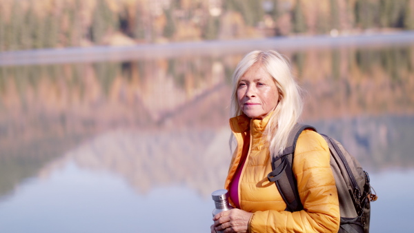 Active senior woman hiker standing by lake outdoors in nature, drinking.